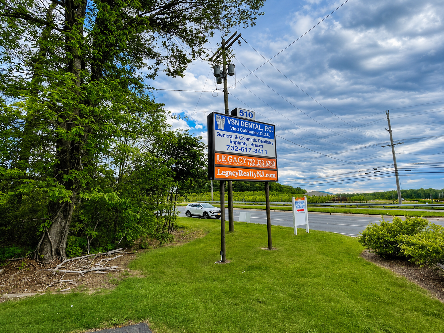 Roadside sign beside a grassy field under a cloudy blue sky, with a paved road and trees nearby.