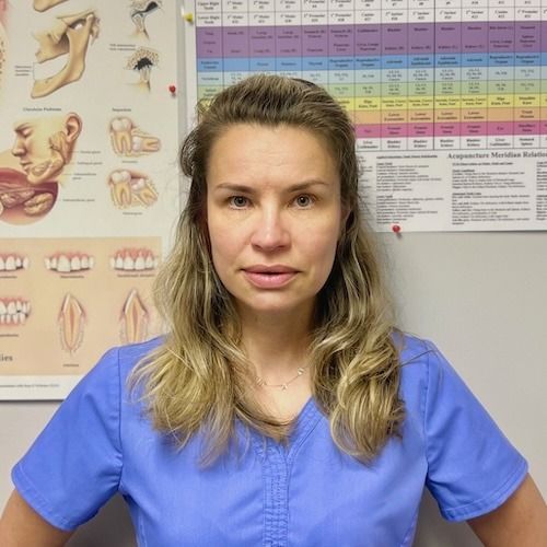 Woman in blue scrubs facing camera in a clinic, with dental anatomy charts on the wall behind her.
