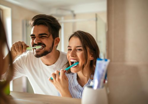 Two people brushing their teeth in a bathroom mirror, smiling with colorful toothbrushes.