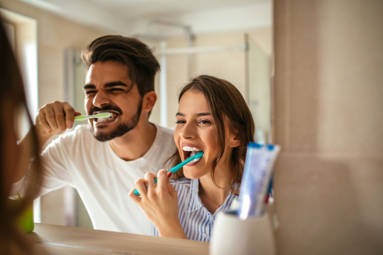 Two people brushing their teeth in a bathroom mirror, smiling with colorful toothbrushes.