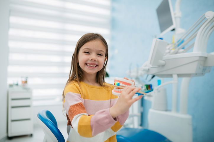 Smiling child holding a toothbrush in a bright dental clinic, seated in a blue chair