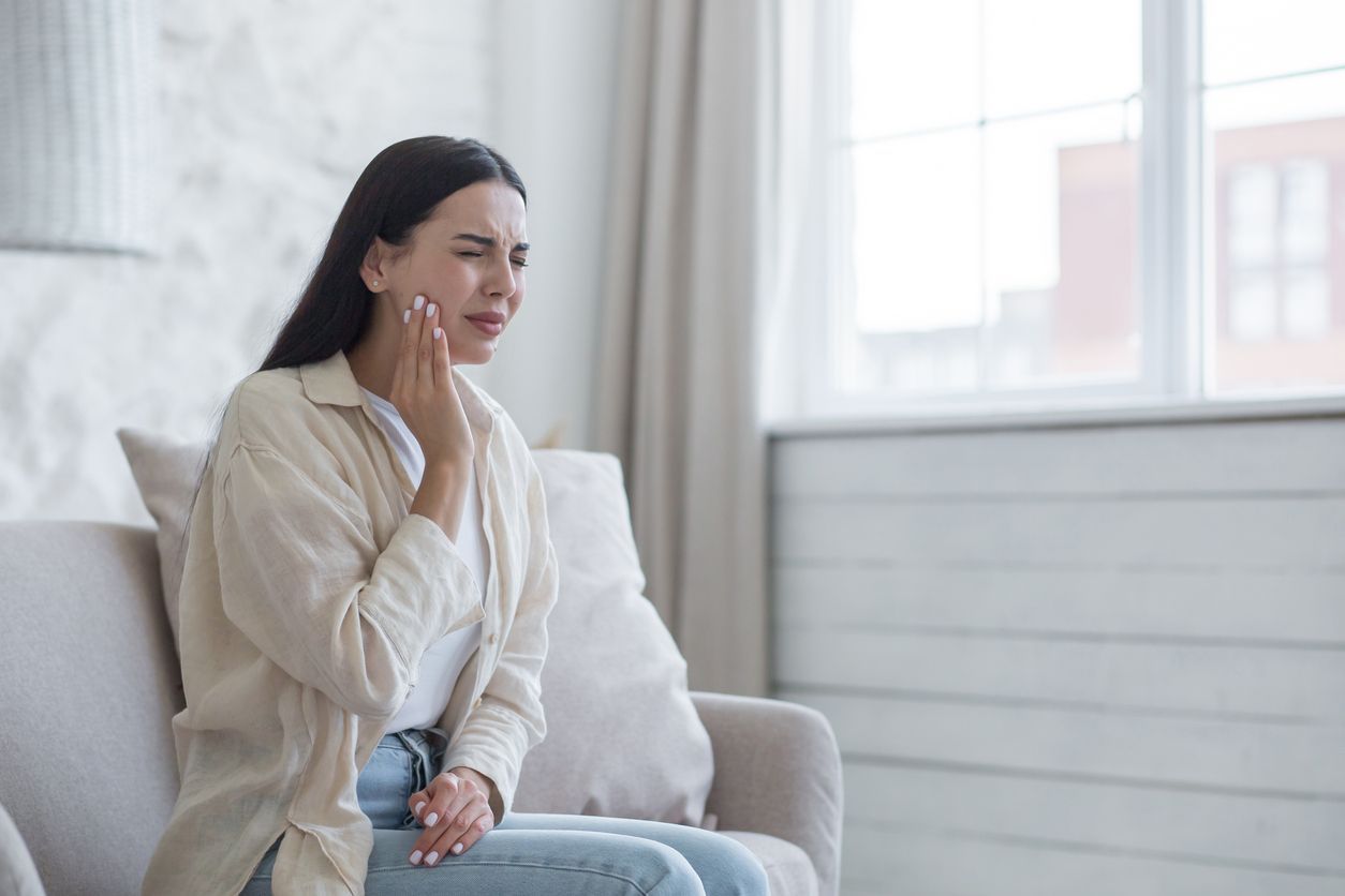 Person holding face with eyes closed, sitting on a couch in a dim room with a plant and lamp nearby.
