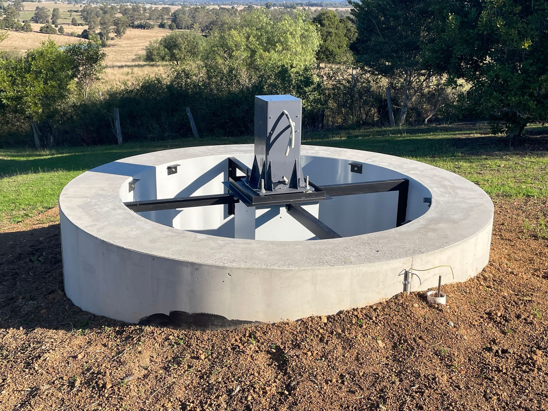 Concrete circular water feature with metal structure, set on a dirt hill overlooking a grassy landscape
