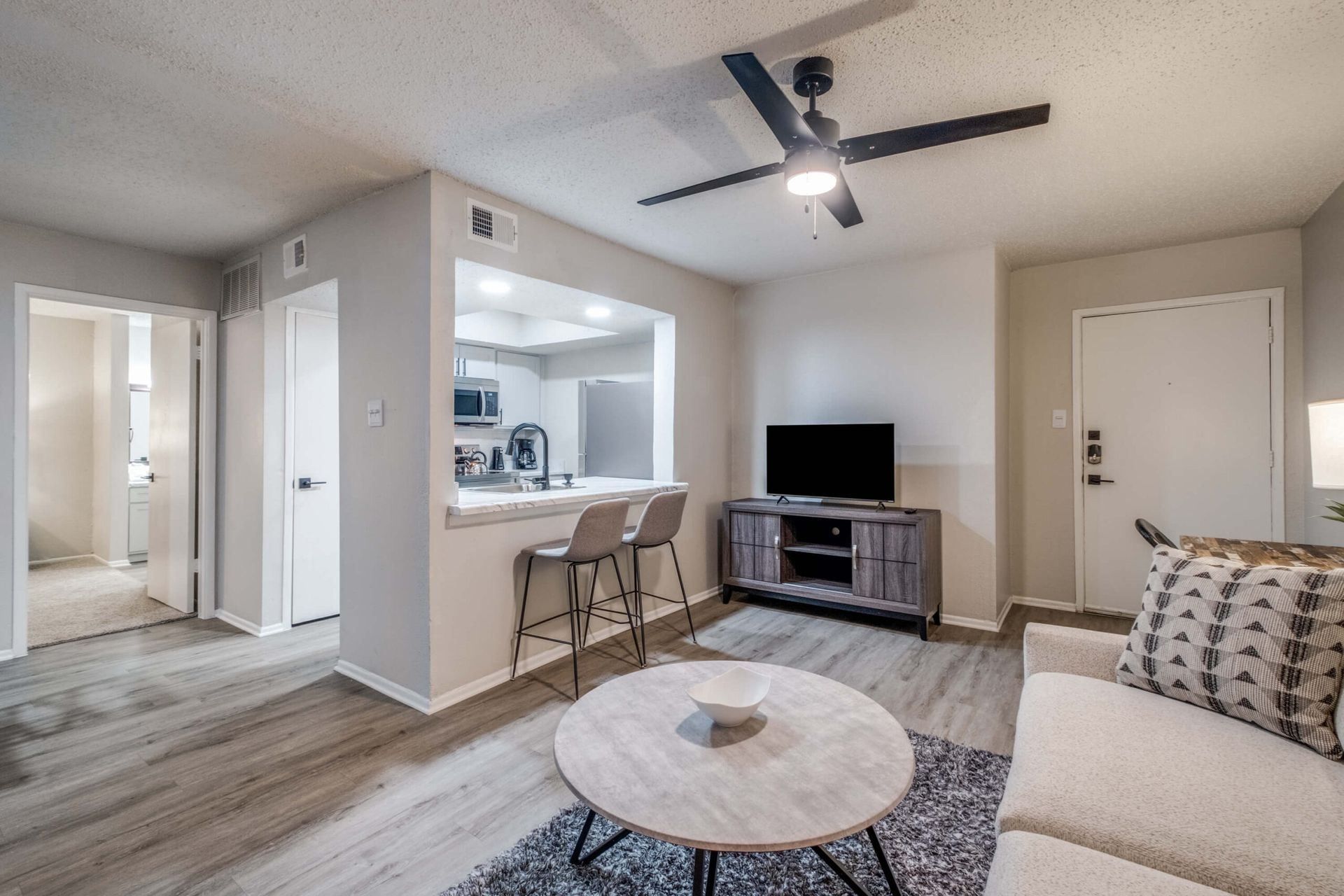 Bright living room with sofa, round coffee table, TV, ceiling fan, and kitchen pass-through with bar stools