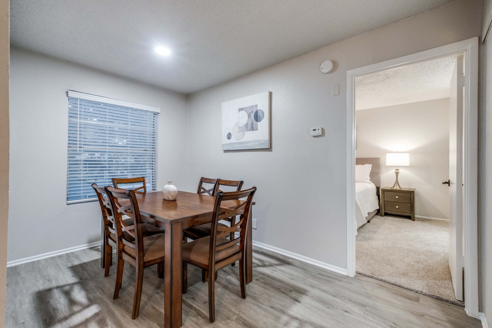 Dining area with wooden table and chairs, light gray walls, and a view into a carpeted bedroom.