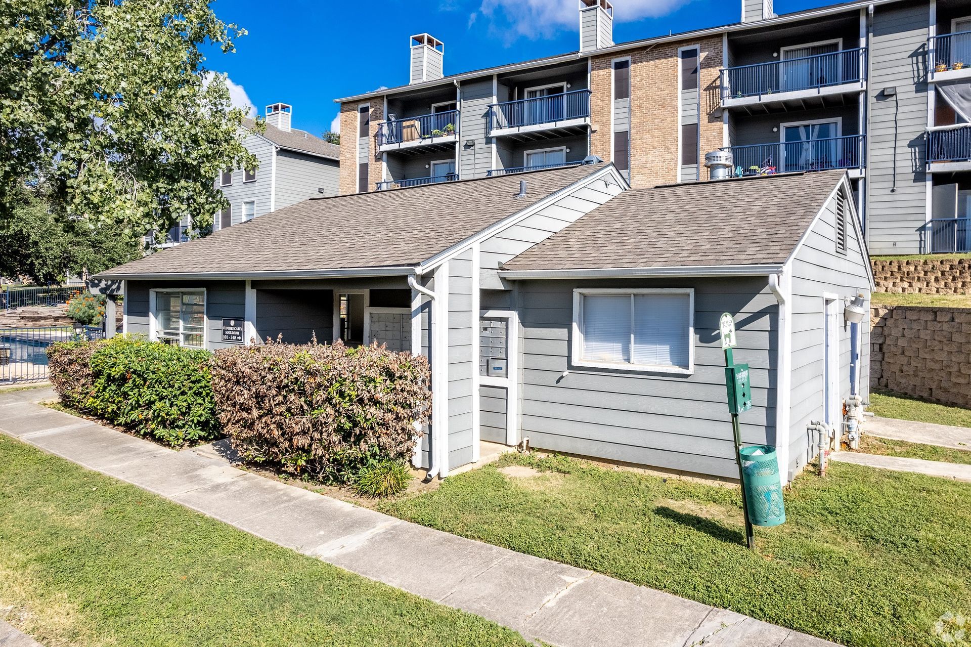 Row of small gray sheds beside an apartment building, with a sidewalk and grass in front.