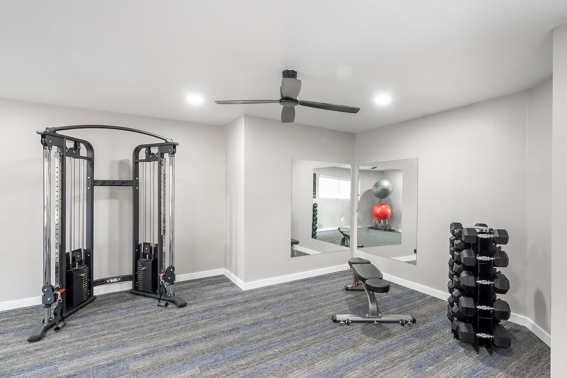 Modern home gym with cable machine, bench, dumbbells, and wall mirror on gray carpet.