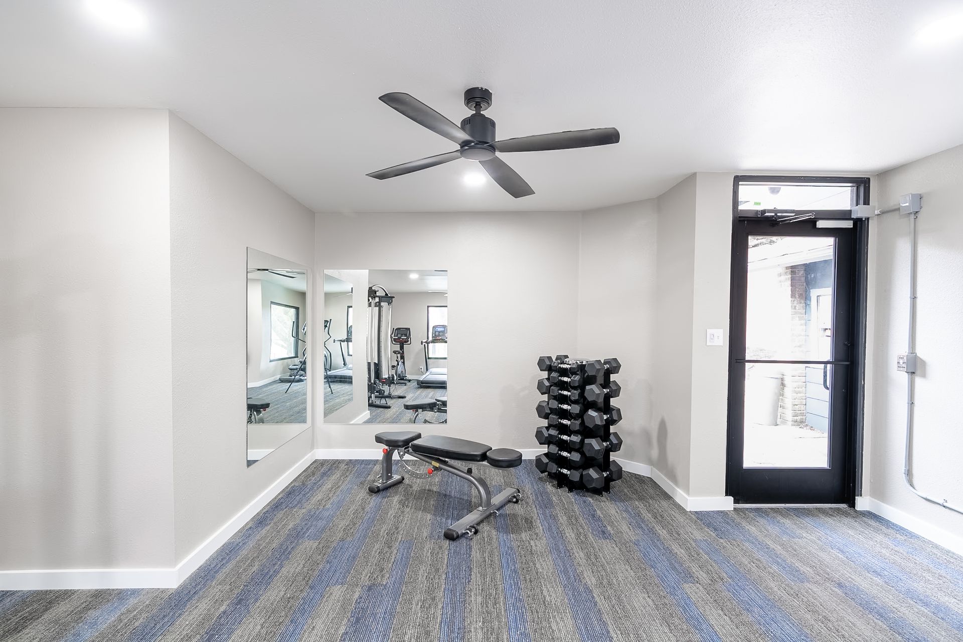 Empty gym room with dumbbell rack, weight bench, mirrors, and ceiling fan near a glass door.