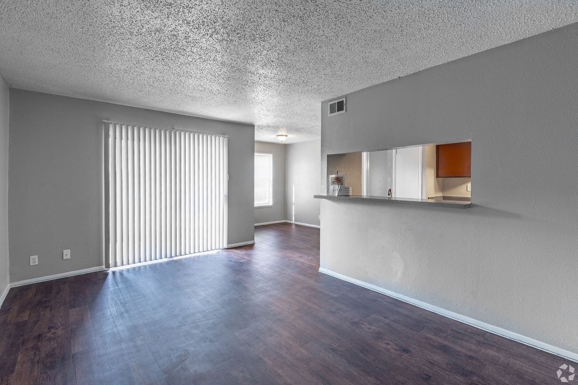 Empty living room with gray walls, dark wood floor, sliding glass door, and pass-through kitchen counter.