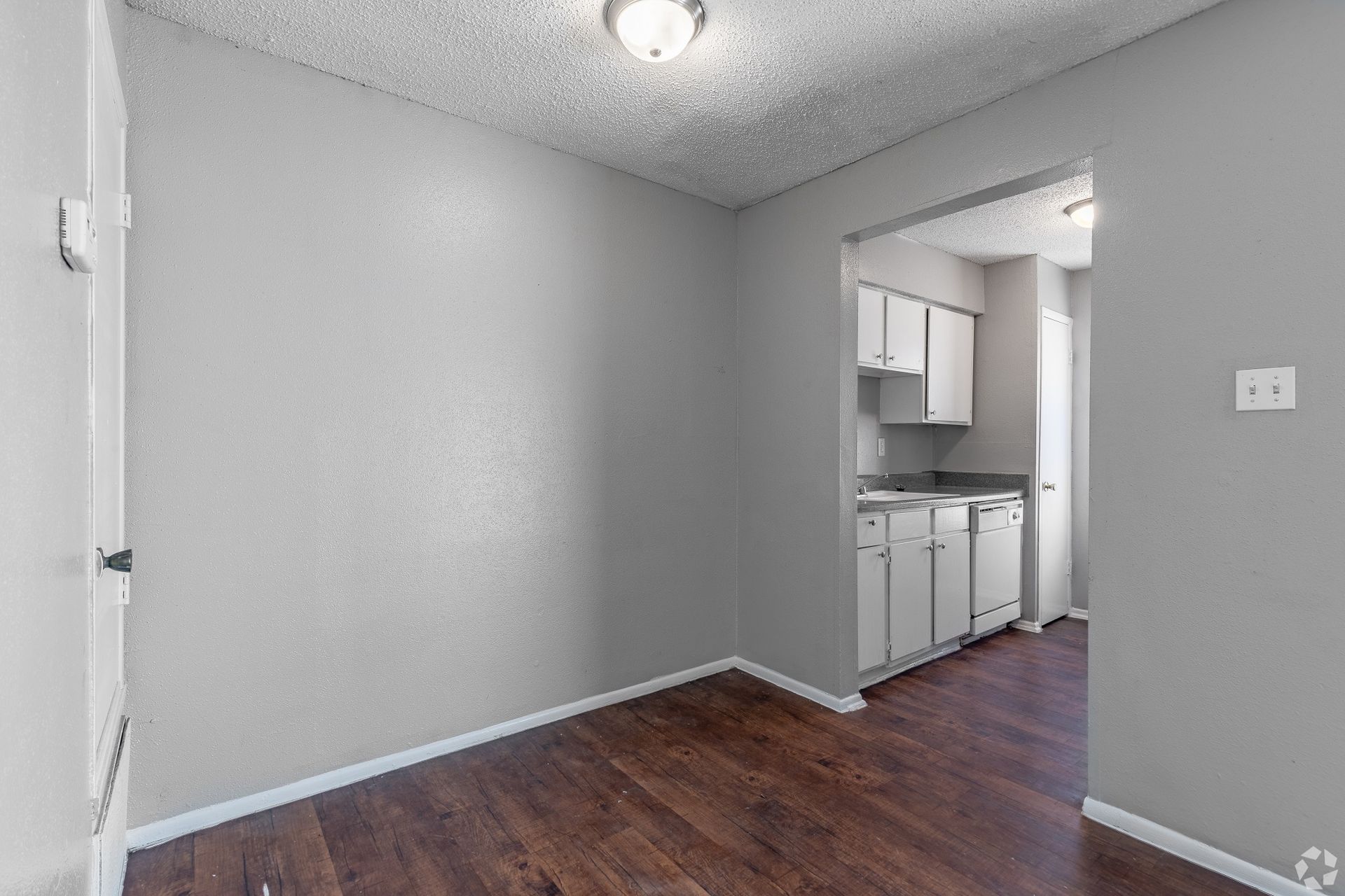 Empty gray room with wood floor and a view into a small white kitchen