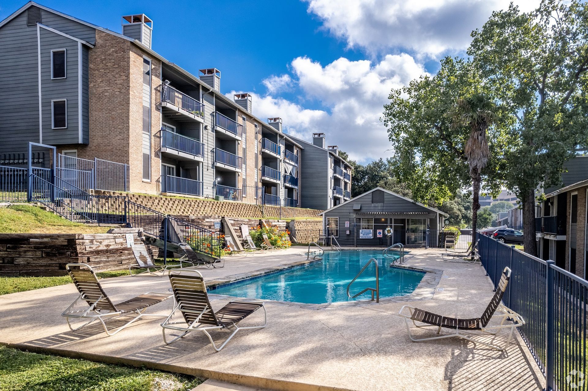 Apartment complex pool with lounge chairs and a blue sky behind trees