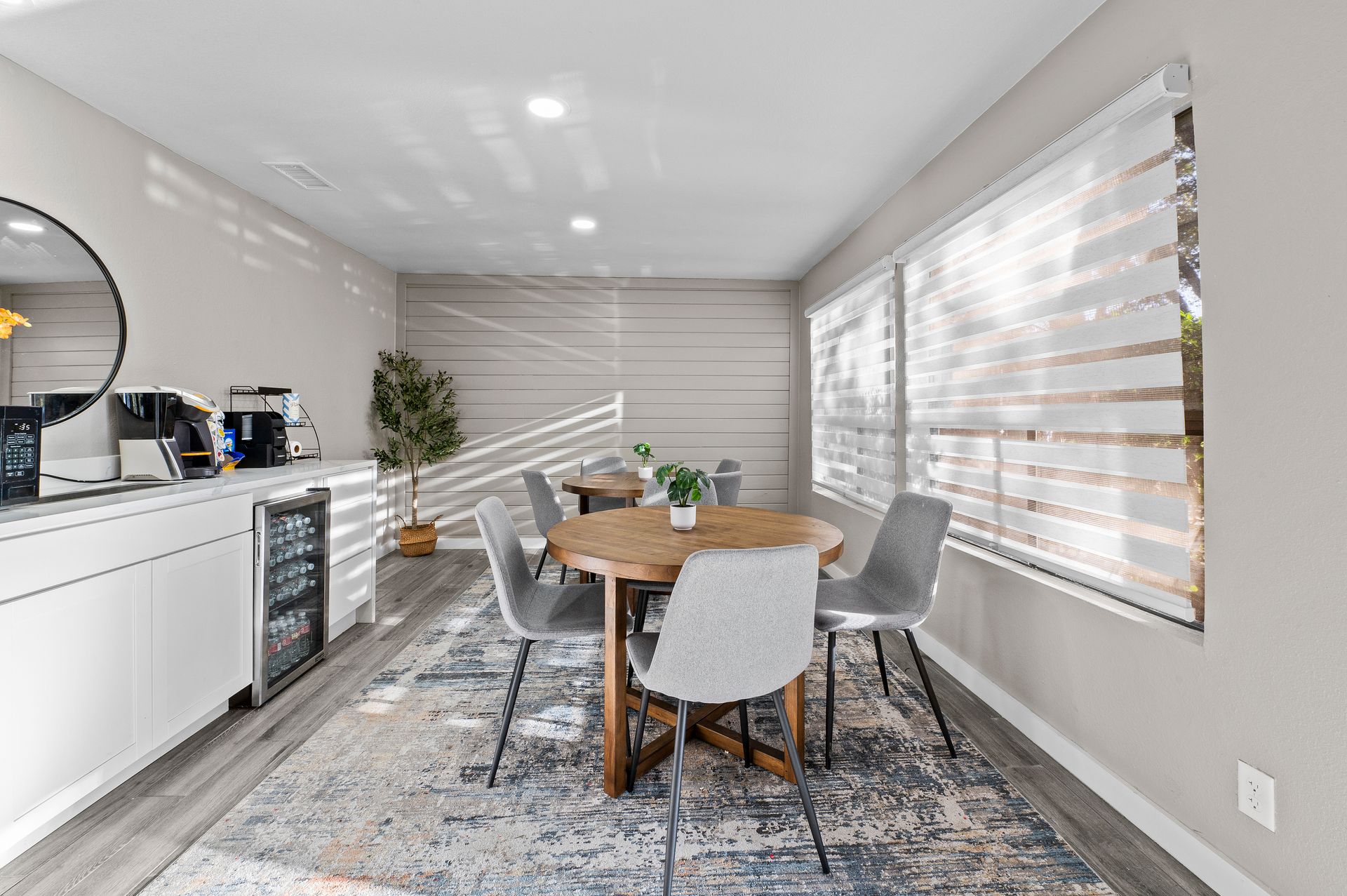 Modern dining area with round wooden table, gray chairs, white cabinets, and large window blinds