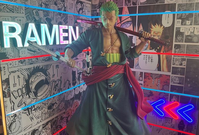 Statua di spadaccino in stile anime con tunica verde, insegna al neon di un ristorante ramen e decorazione murale in stile fumetto.