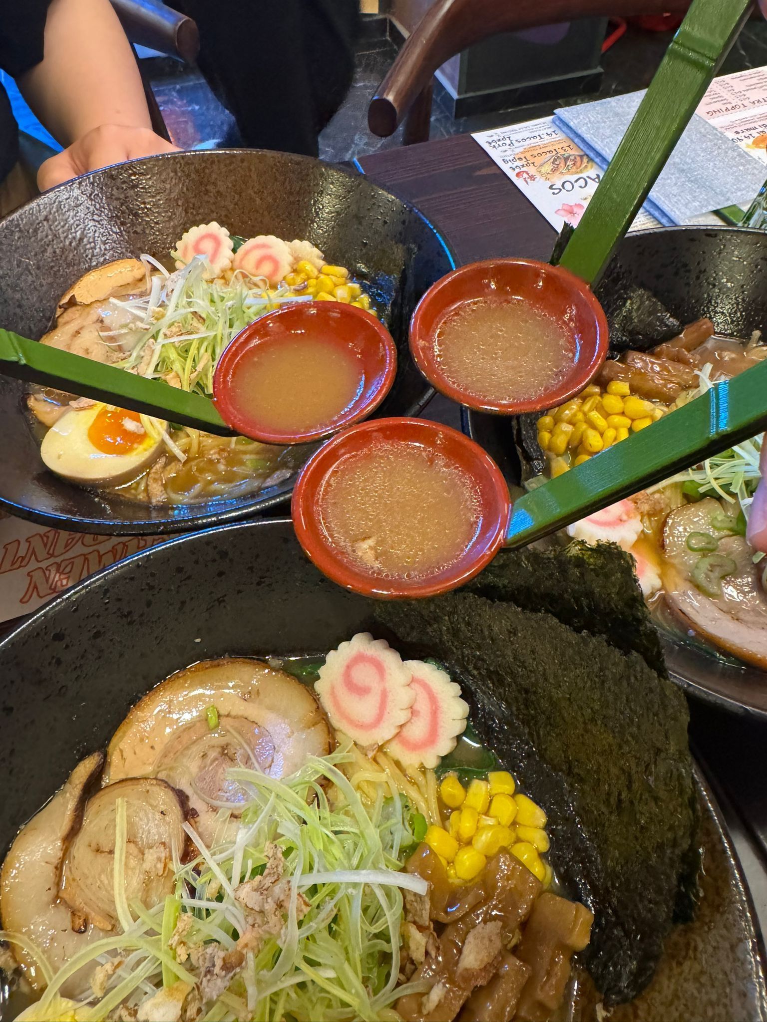 Su un tavolo, ciotole di ramen con mestoli rossi, guarnite con uovo, mais e tortino di pesce.