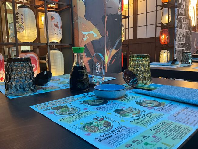 Tavolo da sushi bar con menù, bacchette, piattini e lanterne luminose sullo sfondo.