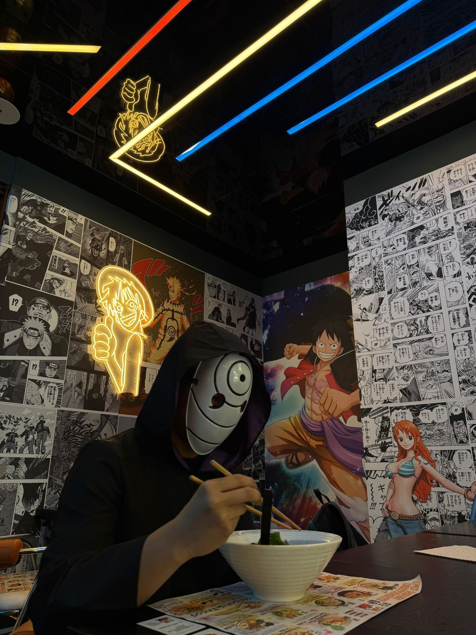 Una persona con indosso una maschera da cartone animato mangia spaghetti in un tavolo di un ristorante illuminato al neon.