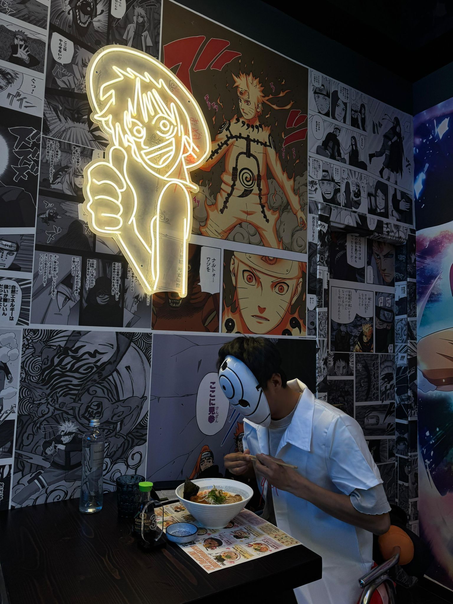 Una persona pranza a un tavolino in un caffè a tema anime illuminato al neon con pareti decorate con collage.