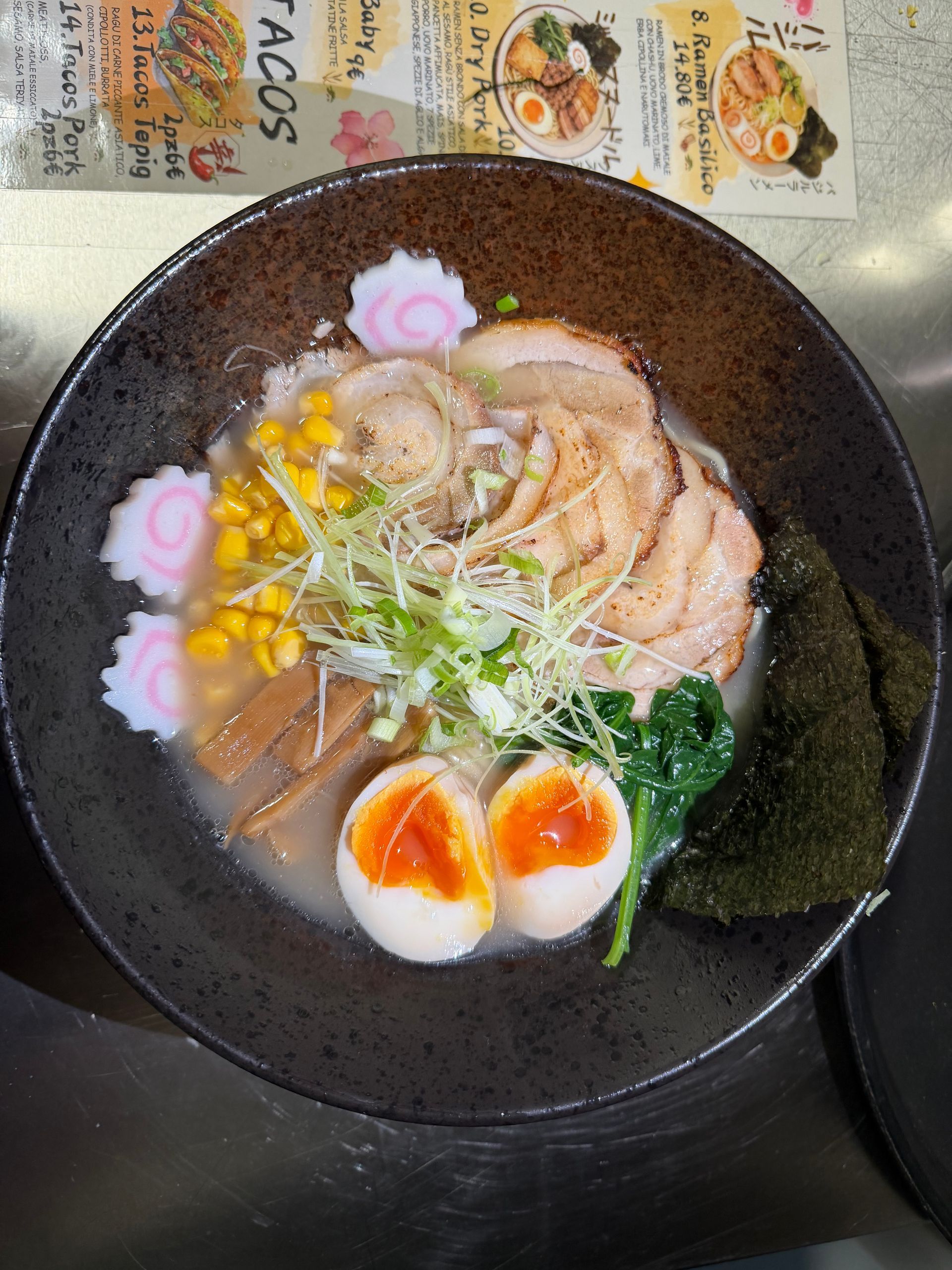 Una ciotola di ramen con carne di maiale a fette, uova alla coque, mais, verdure e naruto (polpette di pesce) su un tavolo scuro.