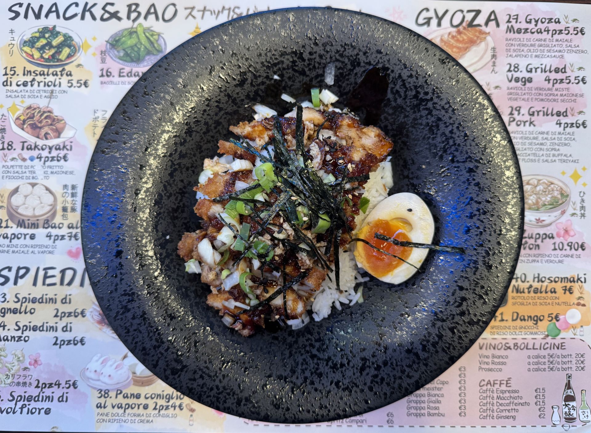 Una ciotola nera di riso condita con salsa, erbe aromatiche e un uovo alla coque, sullo sfondo di un menù di ristorante.