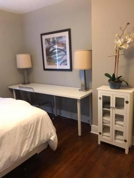 Bedroom with white bed, gray walls, two lamps, framed art, and a small white cabinet with flowers.