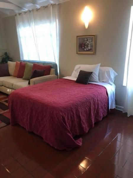 Bedroom with a red bedspread, cream sofa, and wall art beside a bright window.