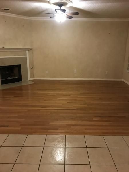 Empty living room with beige walls, hardwood floor, tiled foreground, fireplace, and ceiling fan lights.
