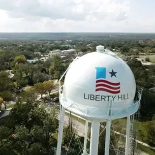 Liberty Hill water tower with Texas flag logo above a wooded neighborhood under cloudy sky