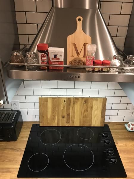 Kitchen stove with black cooktop, metal hood, white tile backsplash, and shelf of spices and jars above.