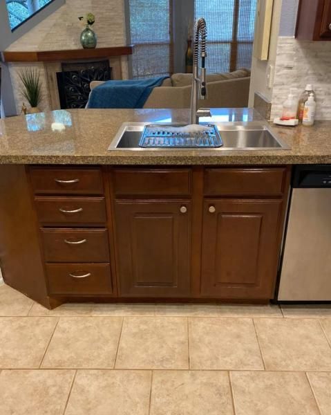 Kitchen sink and dark wood cabinets under granite countertop, with tan tile floor and a dishwasher on the right
