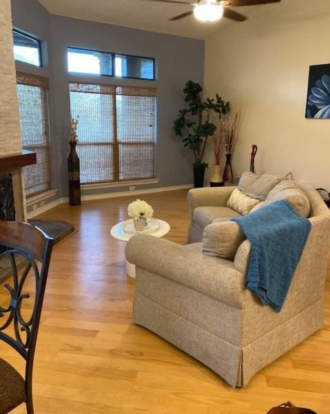 Bright living room with beige armchairs, blue throw, glass coffee table, and large windows with blinds.