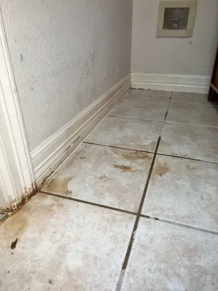 Dirty tiled floor corner with wall stains and a small recessed vent.