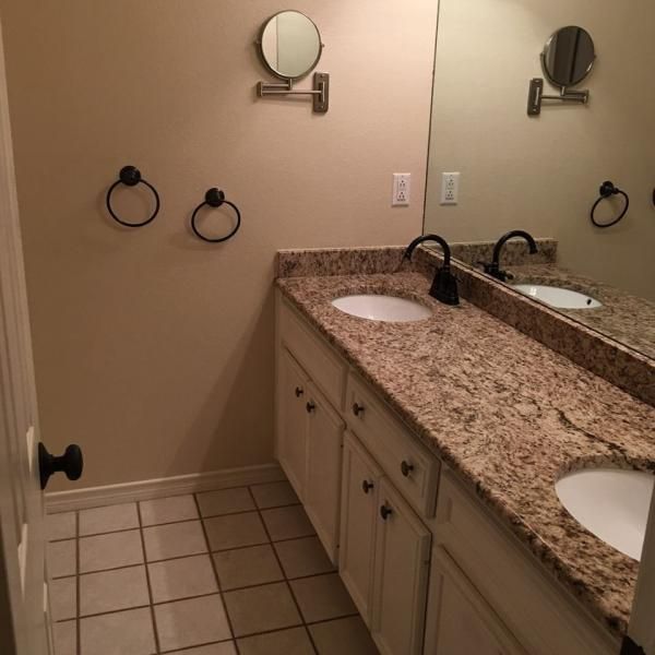Small beige bathroom with granite vanity, double sinks, round mirrors, and tile floor.