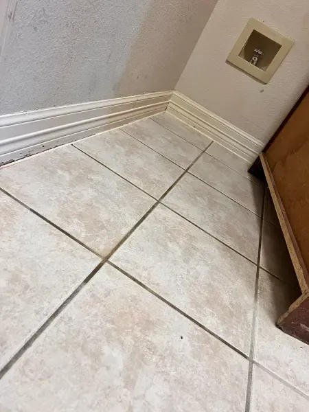 Corner of a tiled floor beside a wall, baseboard, and wooden cabinet.