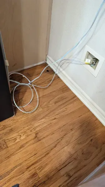 Loose white cables on a wood floor beside a wall outlet and baseboard corner.