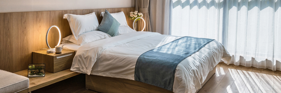 Bright bedroom with a made bed, blue throw blanket, wooden nightstand, and sheer curtains by a large window