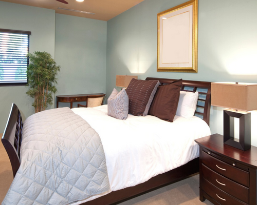 Neatly made bed in a light blue bedroom with nightstands, lamp, window, and framed wall art.