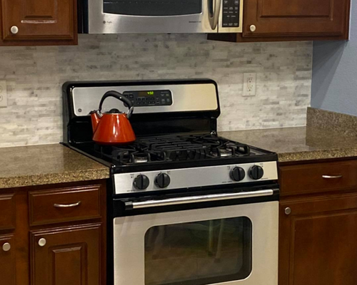 Kitchen stove with orange kettle, stainless oven, wood cabinets, and granite countertops