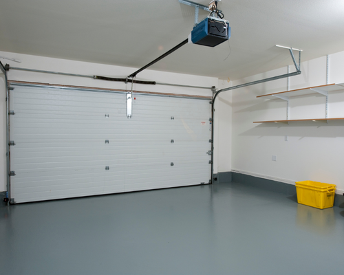 Empty garage with closed white overhead door, gray floor, ceiling opener, shelves, and yellow storage bin.