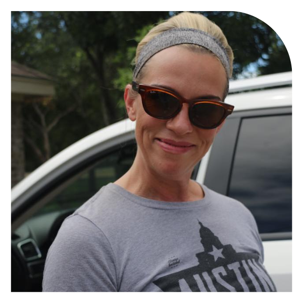 Smiling person wearing sunglasses and a gray headband, standing by a white car outdoors.