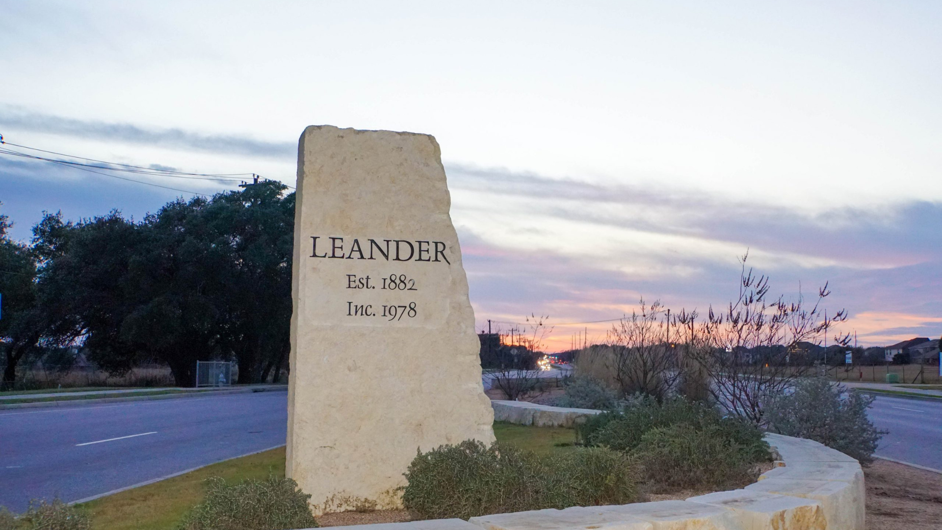 Stone monument by a road at sunset, with “LEANDER” engraved on it.