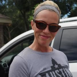 Smiling woman wearing sunglasses and a headband, standing beside a car outdoors