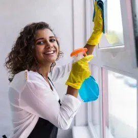 Woman in yellow gloves cleaning a window with a spray bottle, smiling in a bright room