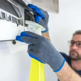 Technician wearing blue gloves cleaning a wall-mounted air conditioner unit with a spray bottle.