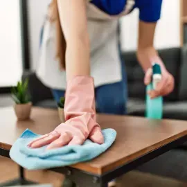 Person cleaning a wooden table with a blue cloth and spray bottle
