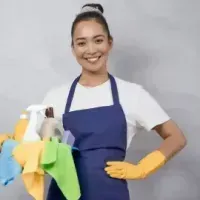 Woman in a blue apron and yellow gloves smiling while holding cleaning supplies with cloths and spray bottles