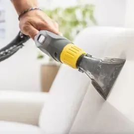 Handheld upholstery cleaner spraying white sofa cushion in a bright living room