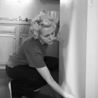 Person crouching and cleaning a wall in a kitchen, with cabinets in the background.