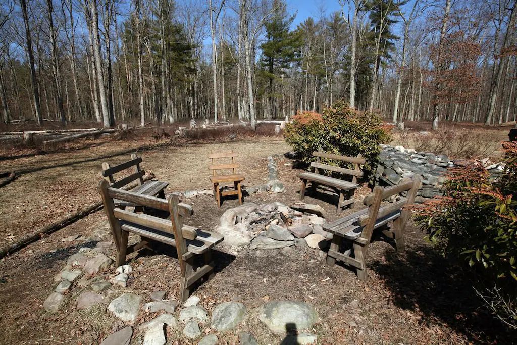 There is a fire pit in the middle of the woods surrounded by wooden benches.