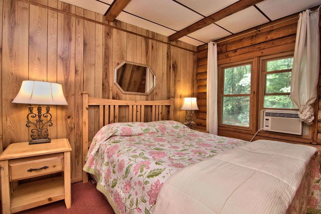 A bedroom in a log cabin with a bed , nightstand , lamp and window.