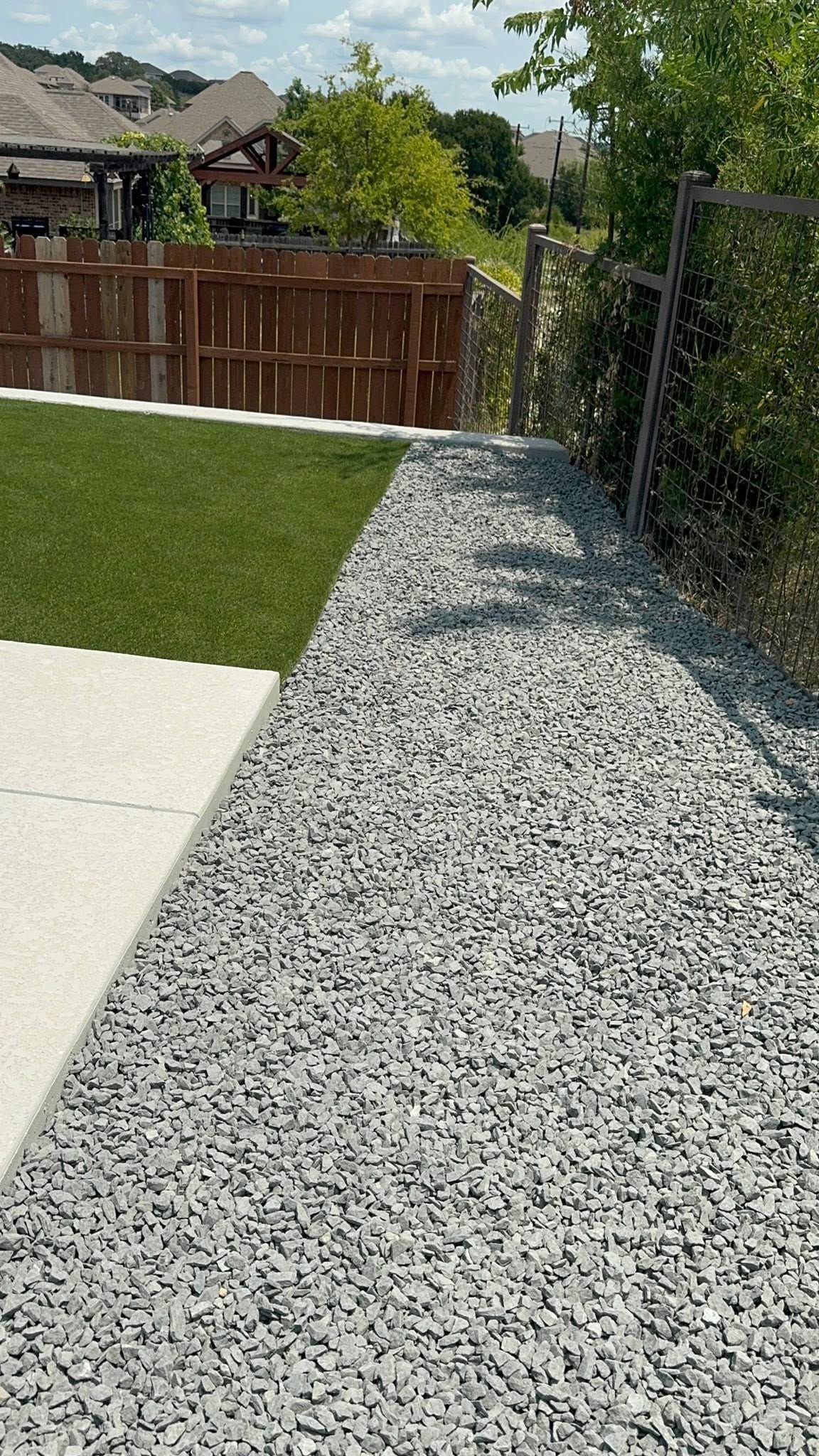 Gravel side yard beside a lawn, with a wooden fence and trees in the background.